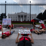 Protestors laying on the pavement in front of the white house holding signs. One sign reads " We deserve better"