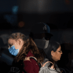 Two women, one wearing a blue disposable mask, the other unmasked.