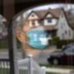 Masked woman photographed through glass with porch, neighborhood houses, and cars reflected.